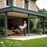 Véranda moderne en aluminium et verre adossée à une maison en pierre bretonne avec une femme examinant des plans d'architecte